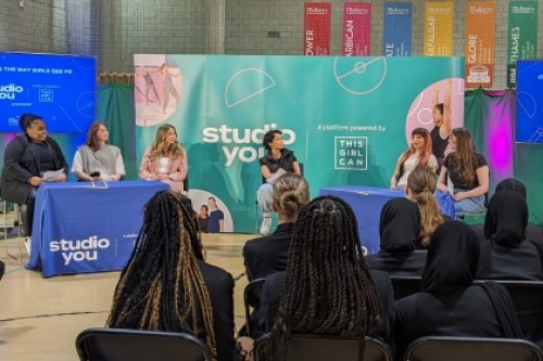 A group of secondary school students sat on chairs in a sports hall watch a panel discussion between a host and five women.